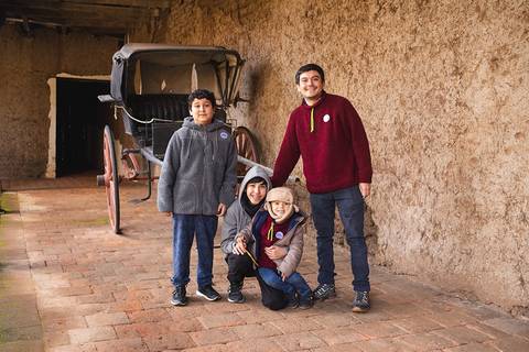 Cuatro niños y un adulto posan sonrientes frente a una carroza antigua en un lugar con paredes de barro.