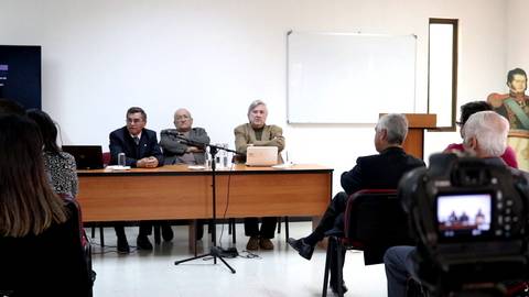 Imagen de un evento académico con varios ponentes en una mesa y un público asistente.