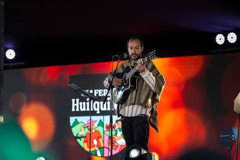 Un músico vestido con un poncho toca la guitarra en un escenario iluminado durante un evento.
