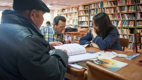 Tres personas están sentadas en una mesa de estudio en una biblioteca, revisando documentos y libros.
