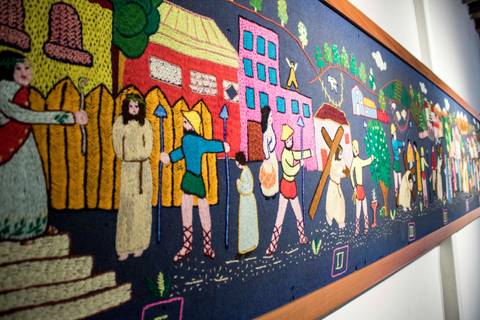 La imagen muestra un mural bordado que representa una escena histórica con personajes y elementos de un pueblo.