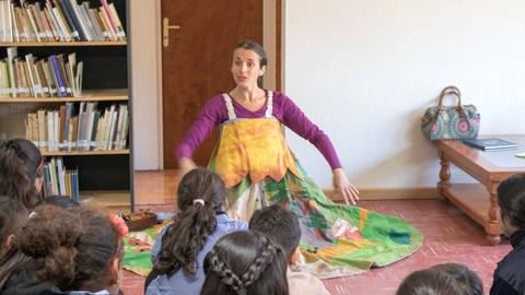 Una narradora de cuentos se presenta ante un grupo de niños, usando un vestido colorido que parece un paisaje.