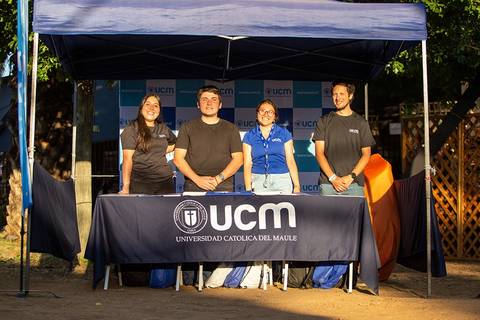 Un grupo de cuatro personas se encuentra de pie frente a una mesa con el logotipo de la Universidad Católica del Maule bajo una carpa.