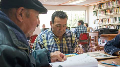 Un grupo de hombres mayores está sentado en una biblioteca, concentrados en leer y discutir sobre libros.
