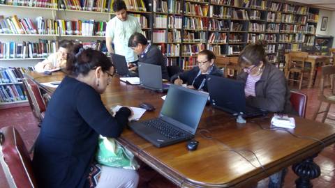 Un grupo de personas trabajando en una sala de estudio con laptops y estanterías de libros de fondo.