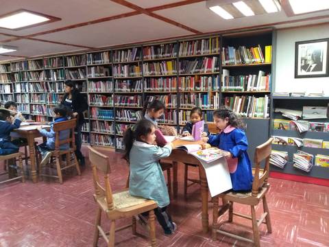 Un grupo de niños está leyendo y dibujando en una biblioteca con estanterías repletas de libros.