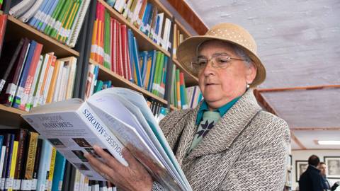 Una mujer mayor lee un libro en una biblioteca, rodeada de estantes llenos de libros.