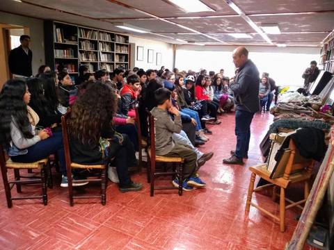 Un grupo de estudiantes escucha atentamente a un orador en un salón con estanterías de libros.