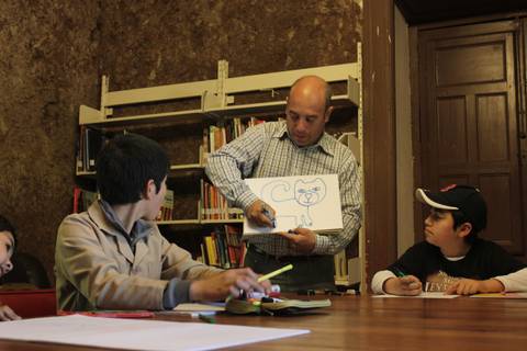 Un hombre dibuja un gato en una hoja mientras varios niños lo observan en una biblioteca.