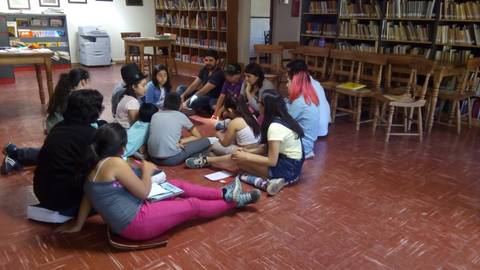 Un grupo de jóvenes está reunido en círculo en una biblioteca mientras comparten ideas y actividades.