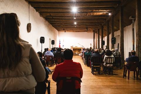 Una reunión con un gran número de personas en un salón con un escenario al frente.