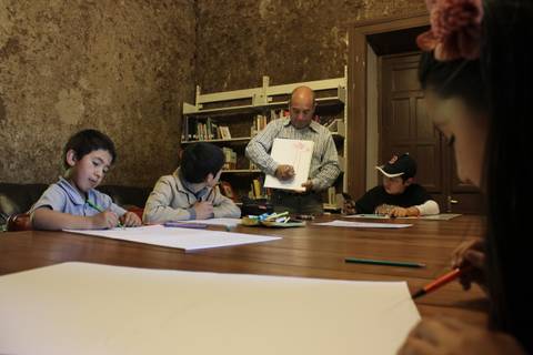 Un grupo de niños dibujando con la supervisión de un adulto en un aula con estanterías de libros.