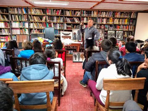 Un hombre está dando una charla frente a un grupo de niños en una biblioteca llena de libros.