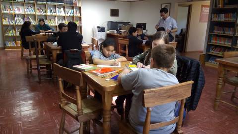 Un grupo de niños y adultos trabajando en un aula con mesas y estanterías de libros.