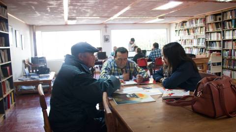 Tres personas se sienten alrededor de una mesa en una biblioteca, conversando y trabajando en un proyecto conjunto.