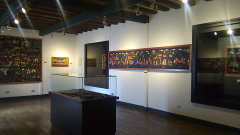 La imagen muestra una galería de arte con obras coloridas en las paredes y una vitrina en el centro.