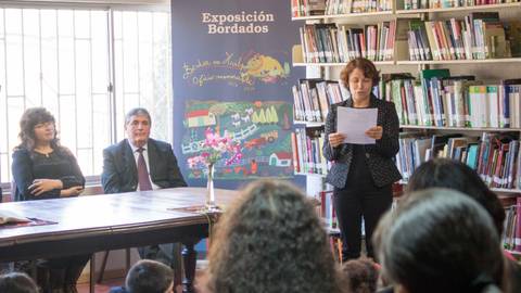 Una mujer lee un texto durante la inauguración de una exposición en una biblioteca, mientras dos personas la escuchan atentamente.