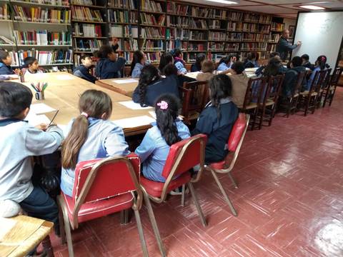 Un grupo de estudiantes está en una biblioteca, sentados en mesas mientras escuchan a un docente.