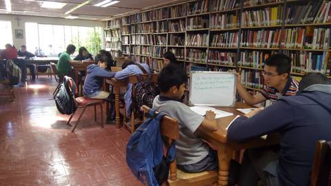 Una biblioteca con varias personas estudiando y un profesor enseñando a un grupo de estudiantes.