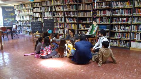 Un grupo de niños y adultos se sienta en el suelo en una biblioteca mientras escuchan una historia.