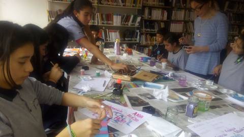 Un grupo de niños y adultos participa en una actividad artística en una biblioteca, pintando y creando manualidades sobre una mesa llena de materiales.