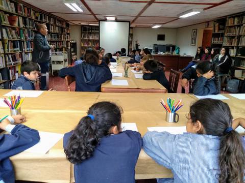 Una clase en una biblioteca donde niños y adultos escuchan a un orador frente a una mesa con material de escritura.