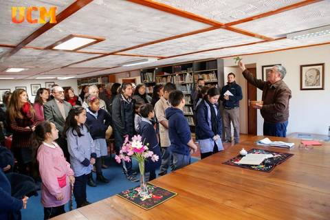 Un grupo de personas escucha atentamente a un hombre que habla y sostiene un objeto en una sala con estanterías llenas de libros.
