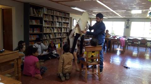 Un grupo de niños se reúne en un salón mientras un adulto sostiene una silla elevada.