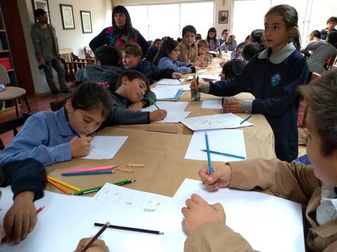 Un grupo de niños sentados en una mesa participan en una actividad de dibujo y pintura en un salón iluminado.