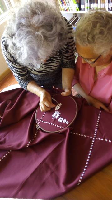 Dos personas mayores están bordando juntas en una tela de color burdeos.