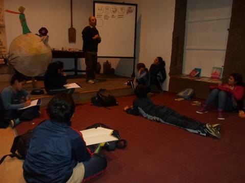 Un grupo de niños escucha atentamente mientras un adulto presenta información en una pizarra en un espacio educativo.