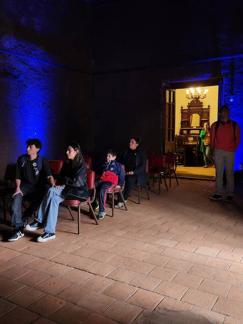 Un grupo de personas sentadas en sillas en una habitación iluminada con luces azules, frente a una puerta que lleva a un ambiente más iluminado.