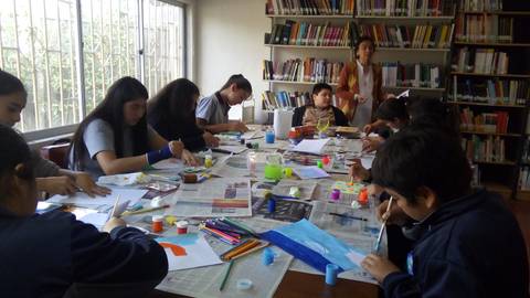 Un grupo de niños y niñas realizan una actividad artística en una mesa llena de materiales creativos en una biblioteca.
