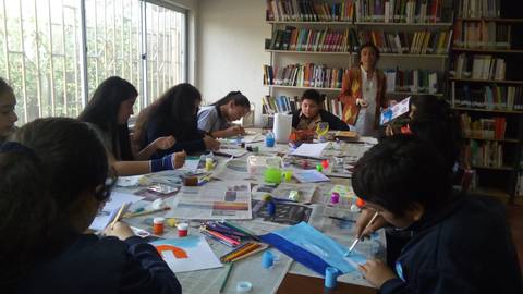 Un grupo de estudiantes está pintando en una mesa llena de materiales artísticos en una sala de clases.