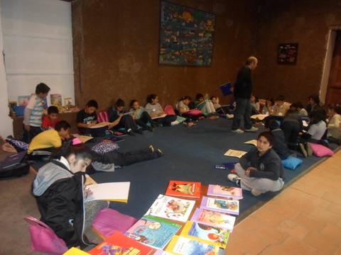 Un grupo de niños y adultos se encuentran en un espacio escolar, leyendo y dibujando en un ambiente relajado.