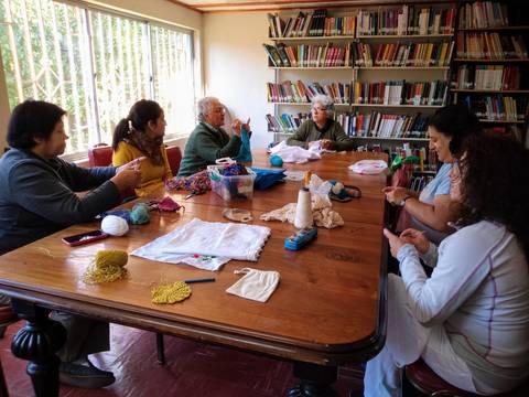 Un grupo de seis personas se reúne en una mesa, realizando actividades de tejido en una sala con estanterías llenas de libros.