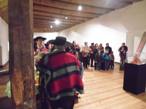 Un grupo de personas observa una exhibición en un espacio cultural mientras un grupo vestido con trajes tradicionales está en el centro.