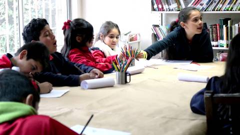 Un grupo de niños se encuentra en una mesa, realizando una actividad creativa mientras una niña habla y señala con la mano.