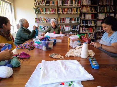Un grupo de mujeres se reúne en una habitación con estanterías llenas de libros, trabajando en manualidades y compartiendo momentos juntas.
