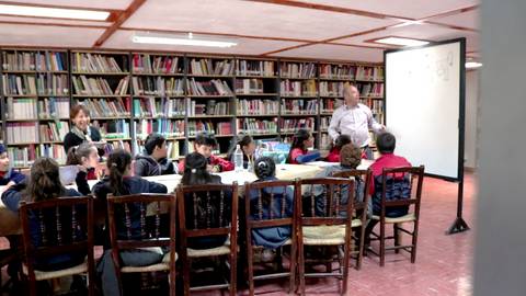 Un maestro enseña a un grupo de niños en una aula frente a una pizarra y rodeados de estanterías llenas de libros.