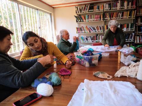 Un grupo de mujeres se dedica a tejer en una mesa rodeada de materiales de trabajo en un ambiente iluminado por la luz natural.