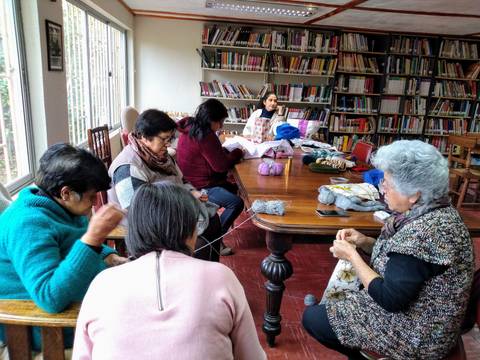 Un grupo de mujeres está tejiendo y compartiendo en una sala con estanterías llenas de libros.