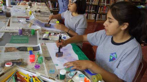 Dos niñas están pintando en una mesa llena de material artístico en un ambiente de biblioteca.