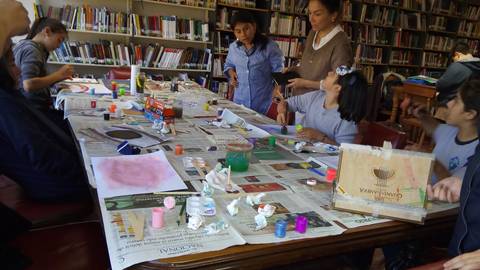 Un grupo de niños y adultos se reúnen en una mesa llena de materiales para una actividad creativa en una biblioteca.