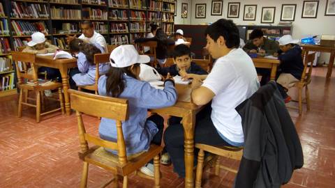 Un grupo de niños y un adulto trabajan en mesas en una biblioteca.