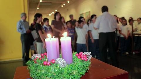 Una escena en una sala donde hay un grupo de personas y en primer plano se observa un centro de mesa con velas moradas y decoración navideña.
