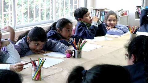 Un grupo de niños sentados en un aula, prestando atención y rodeados de útiles escolares.
