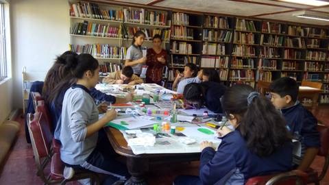 Un grupo de niños están realizando manualidades en una biblioteca, mientras un par de adultos los supervisan.