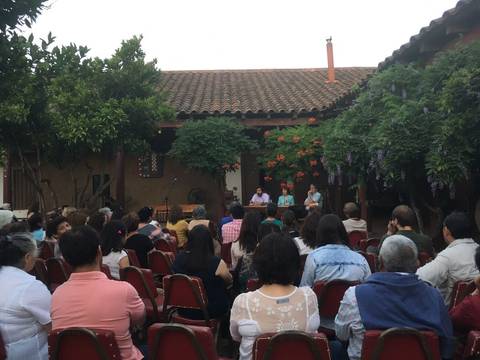 Un grupo de personas se reúne en un patio, sentados frente a un panel que habla en un evento comunitario.
