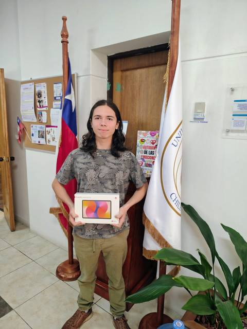 Un joven sosteniendo una tablet frente a una puerta con la bandera chilena y el emblema de la universidad.